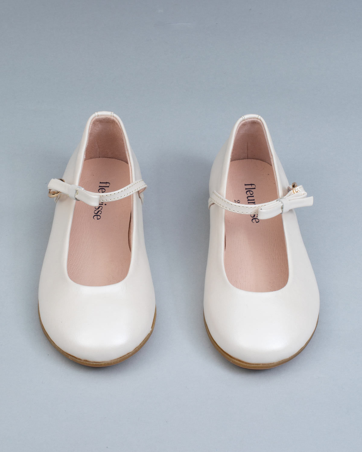 Frontal view of cream leather Mary-Jane shoes for children, showcasing symmetrical bows and rounded toes on a flat background.