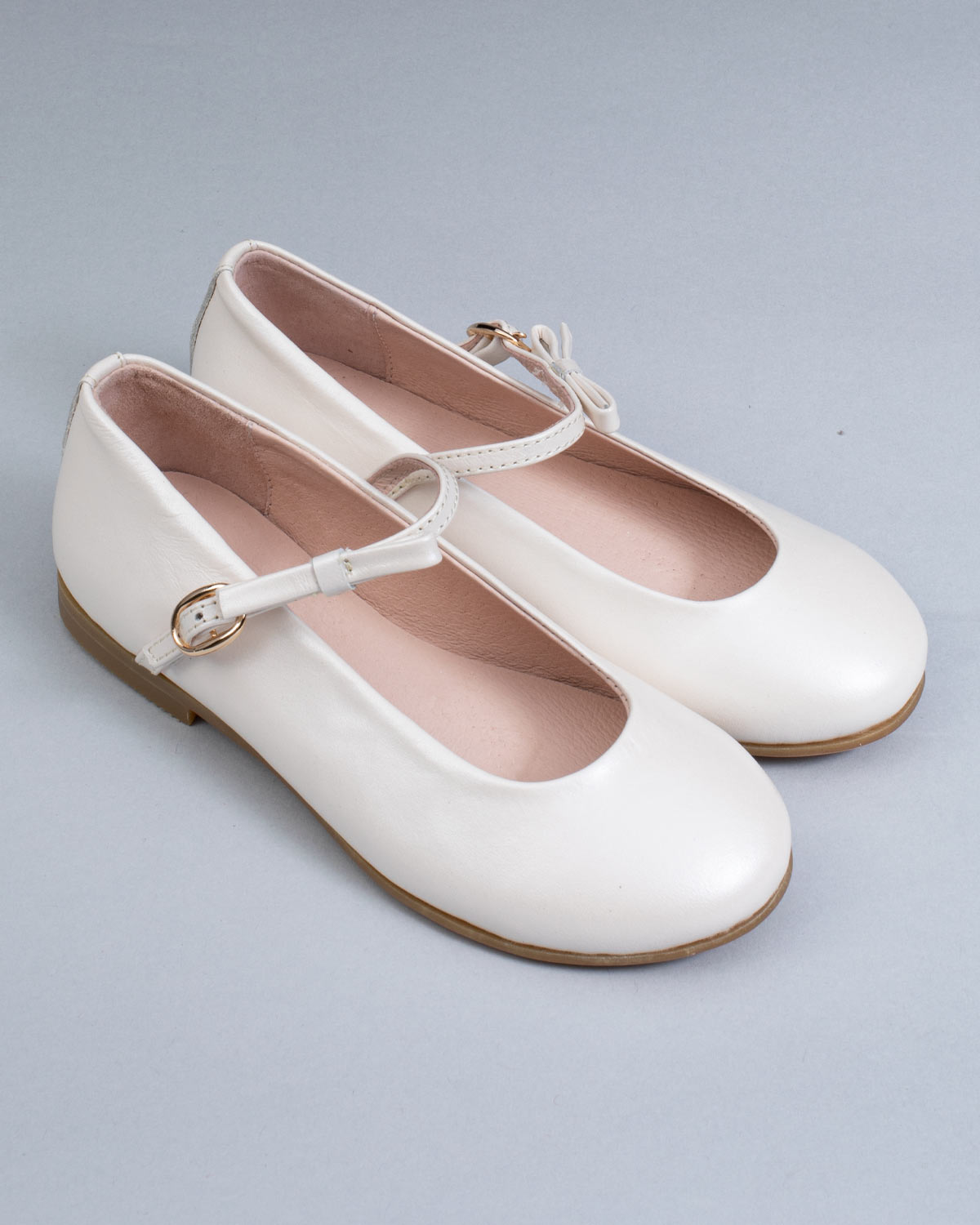 Pair of cream leather Mary-Jane shoes for children, with side buckles and front bows, displayed at an angle on a neutral gray surface.