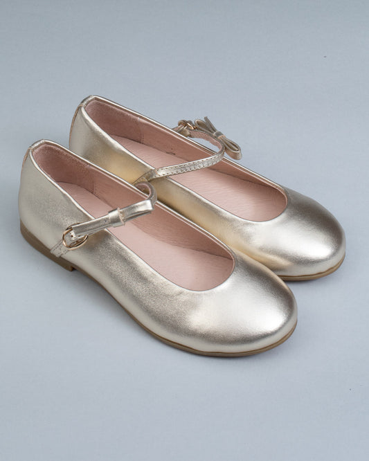 Pair of gold leather Mary-Jane shoes for children, with side buckles and front bows, displayed at an angle on a neutral gray surface.