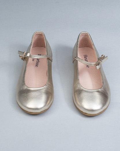 Frontal view of gold leather Mary-Jane shoes for children, showcasing symmetrical bows and rounded toes on a flat background.