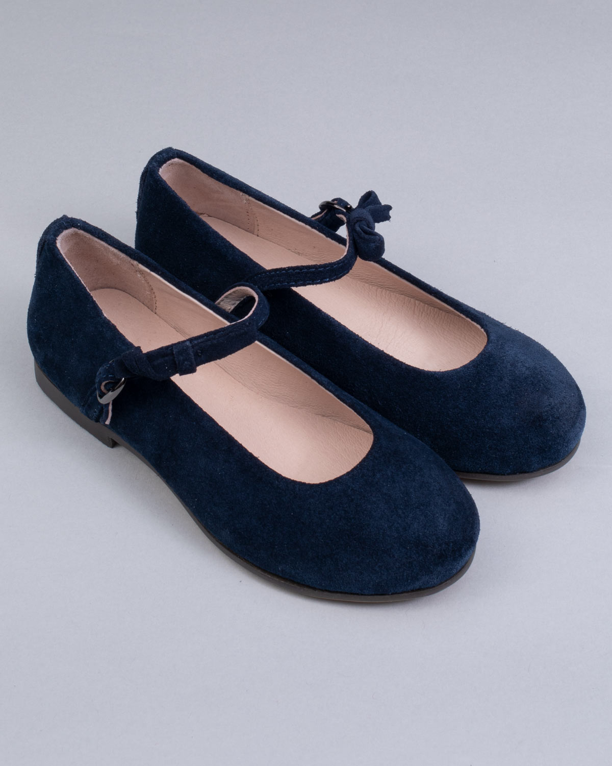 Pair of navy blue suede Mary-Jane shoes for children, with side buckles and front bows, displayed at an angle on a neutral gray surface.