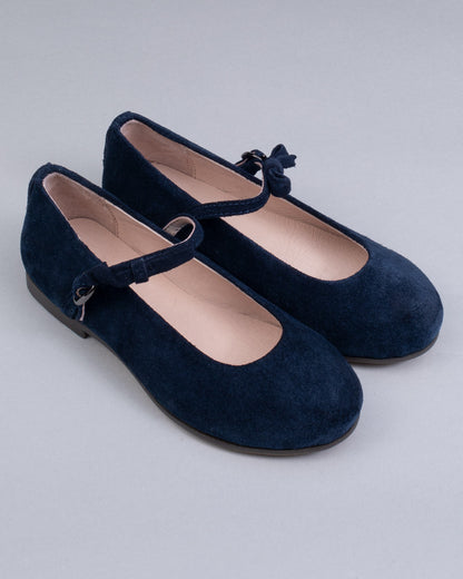 Pair of navy blue suede Mary-Jane shoes for children, with side buckles and front bows, displayed at an angle on a neutral gray surface.