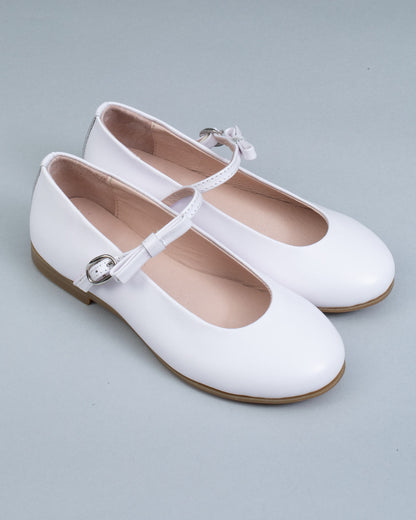 Pair of white leather Mary-Jane shoes for children, with side buckles and front bows, displayed at an angle on a neutral gray 