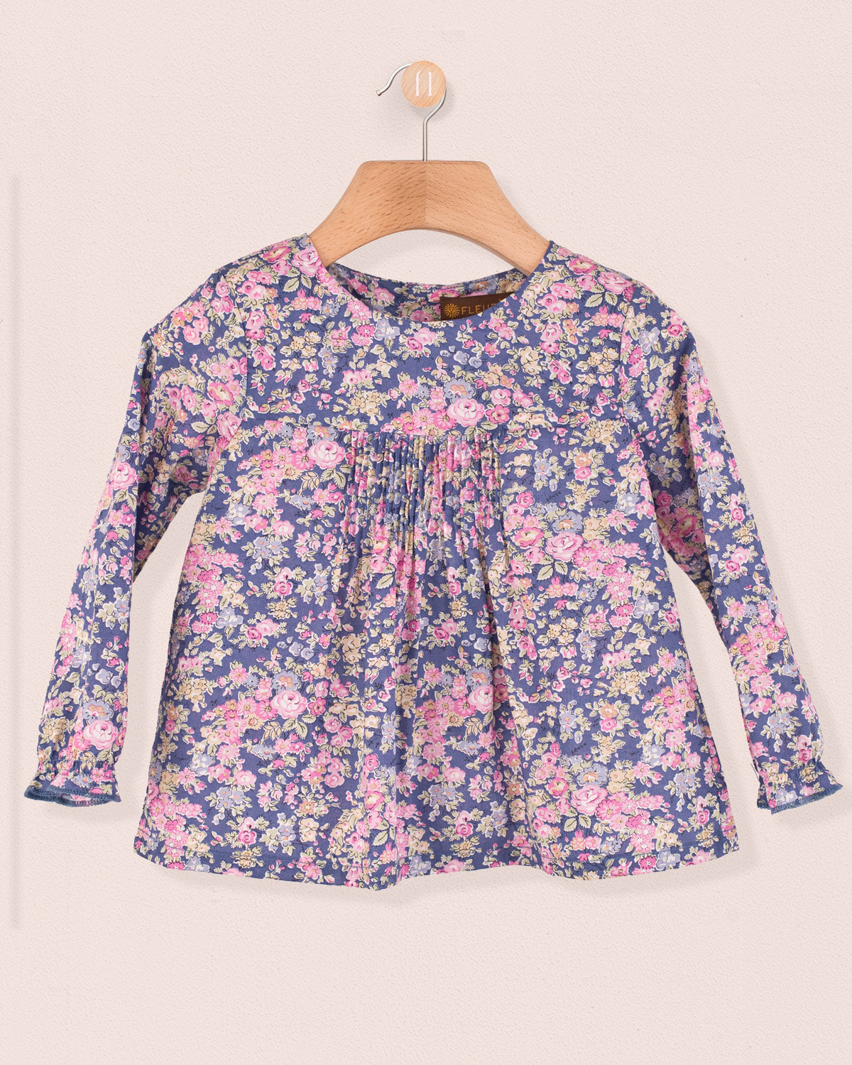 Long-sleeve baby tunic in Liberty Tatum Blue floral cotton with pin-tucks on the chest, displayed on a wooden hanger.