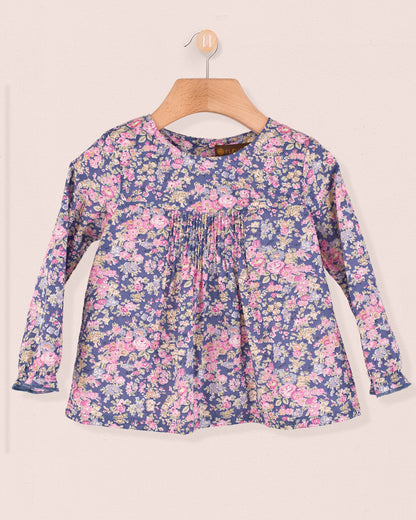 Long-sleeve baby tunic in Liberty Tatum Blue floral cotton with pin-tucks on the chest, displayed on a wooden hanger.