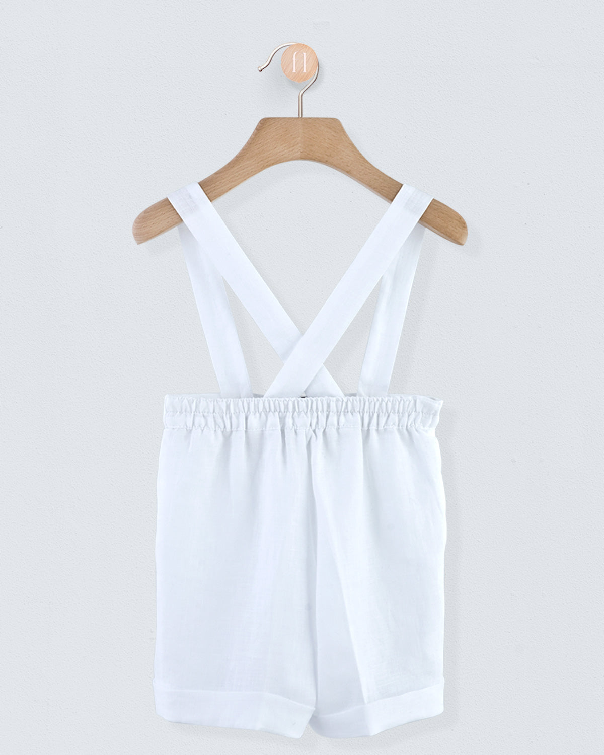 Back view of white linen suspender shorts showing crossed suspenders and gathered waistband, hanging on a wooden hanger