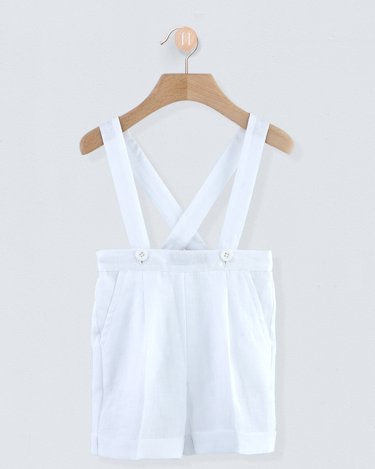 Front view of white linen suspender shorts with corozo buttons, hanging on a wooden hanger