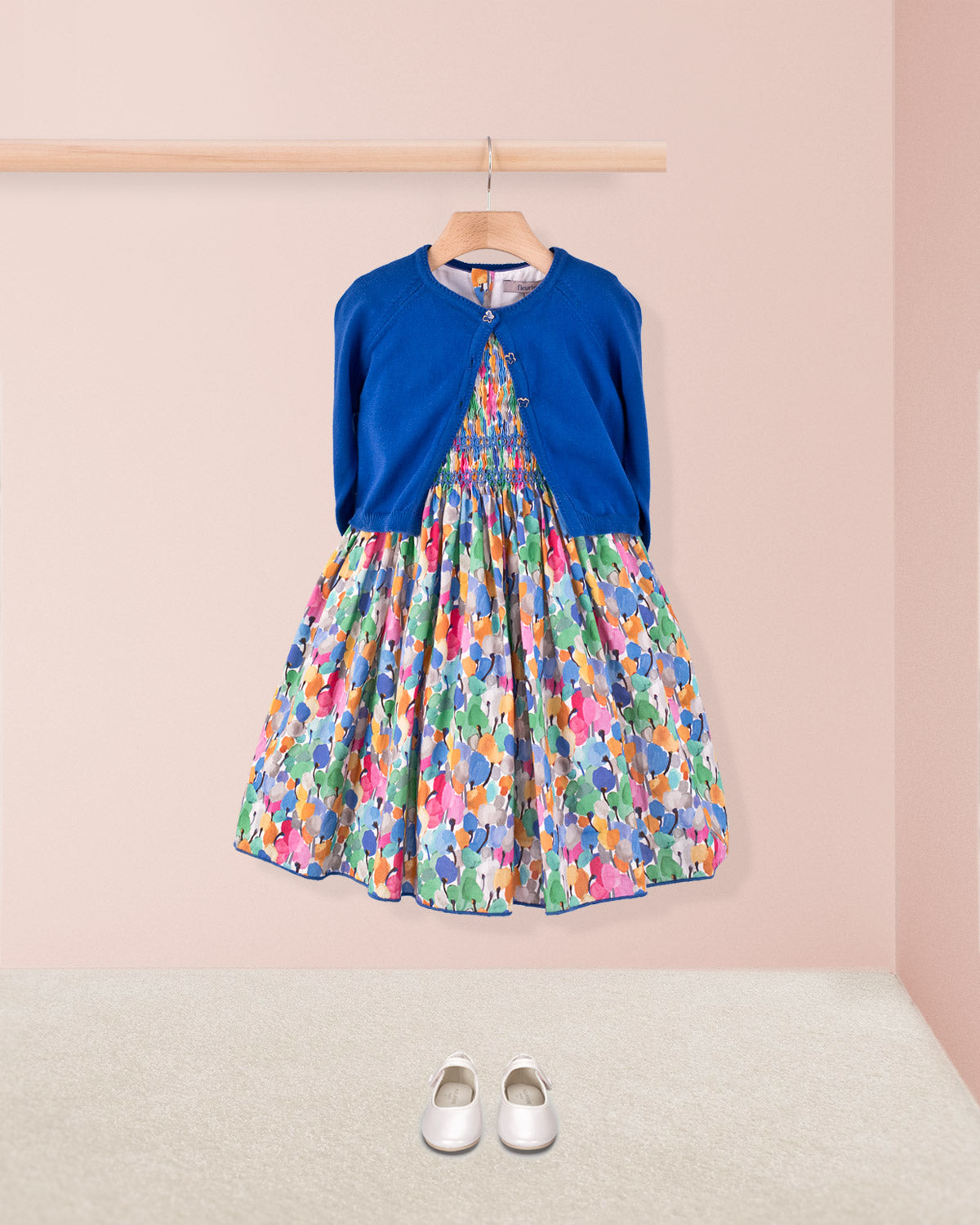 Amandine Liberty Tulip Fields Blue Smock dress styled with a royal blue cardigan and white Mary Jane shoes, displayed against a pale pink background.