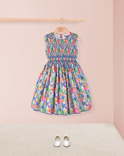 Front view of the Amandine Liberty Tulip Fields Blue Smock dress styled simply with white Mary Jane shoes on a soft carpet, against a light pink wall.