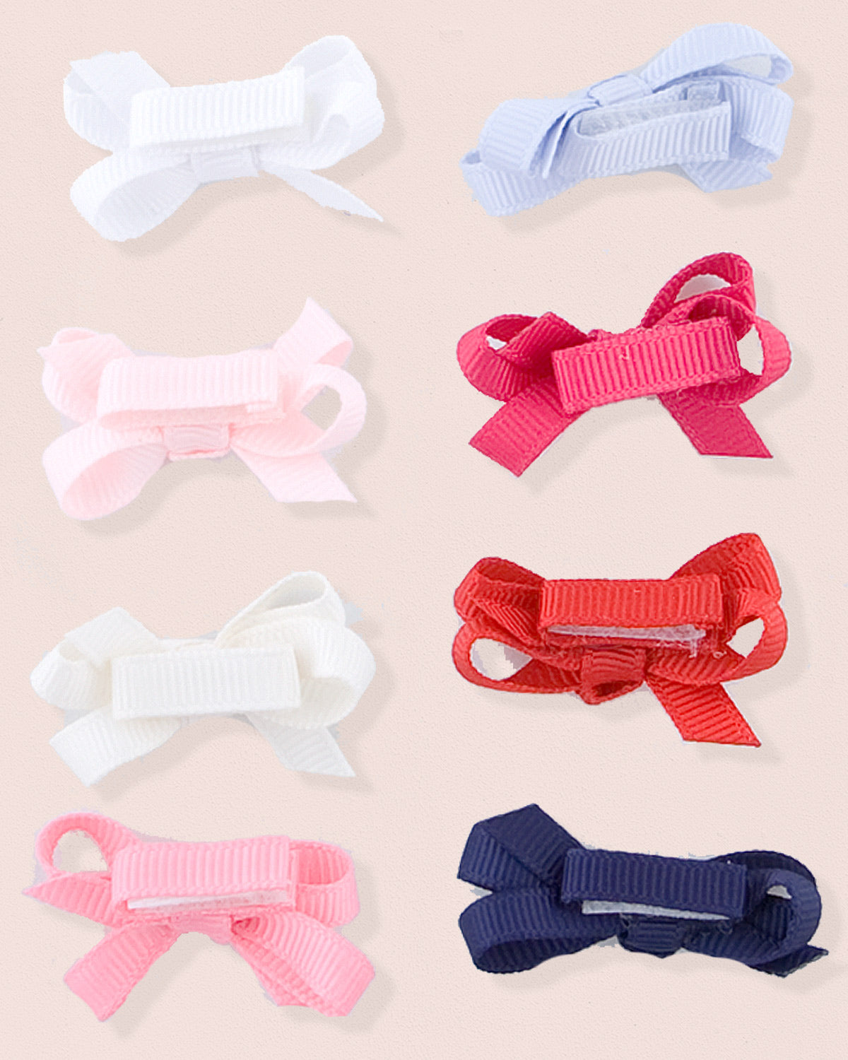 Assortment of grosgrain baby hairbows in various colors including white, light blue, pink, primrose, cream, red, fuchsia, and navy blue, arranged flat on a blush pink background.